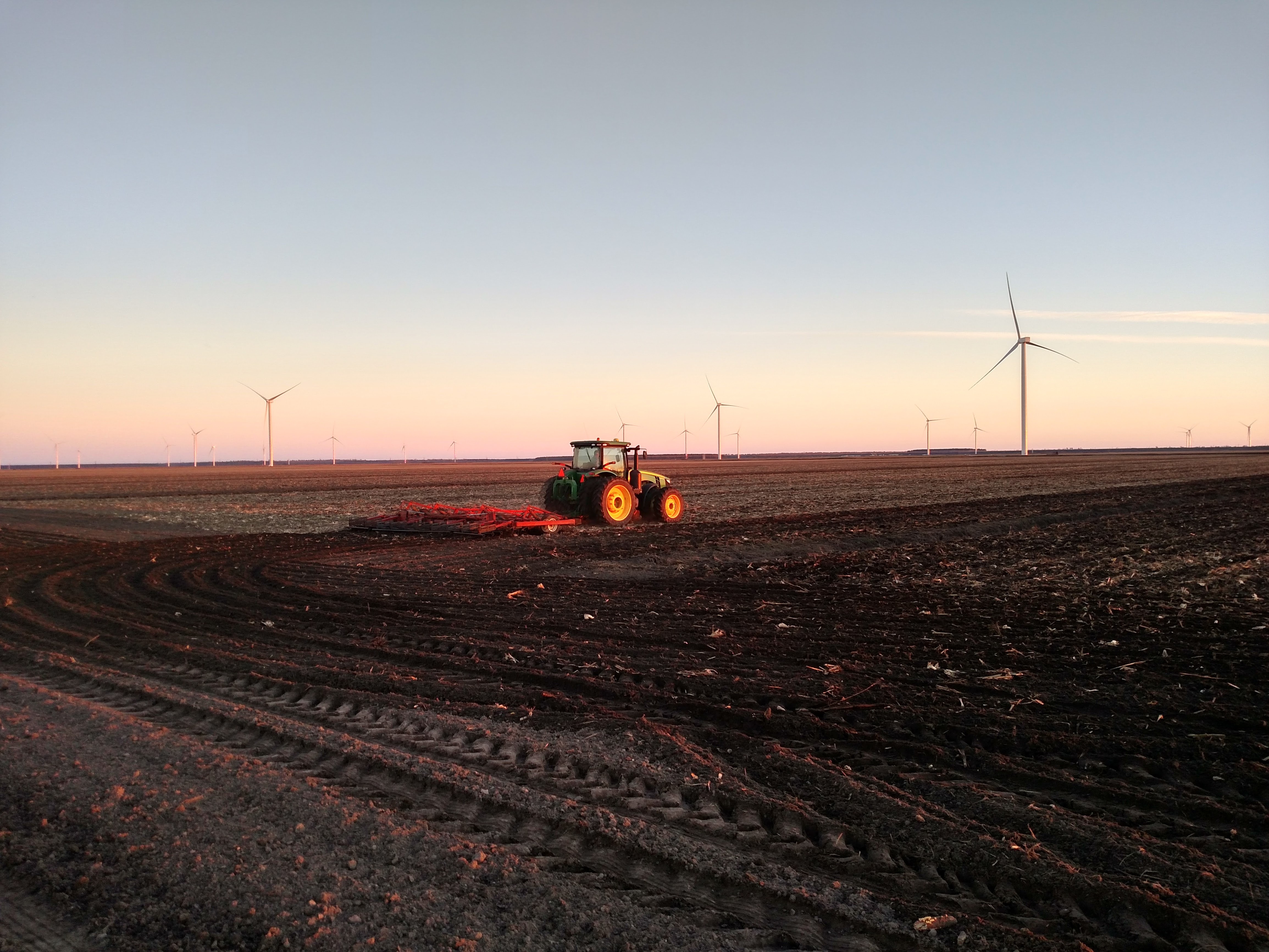 A group of wind turbines behind a plowed field with a tractor in it