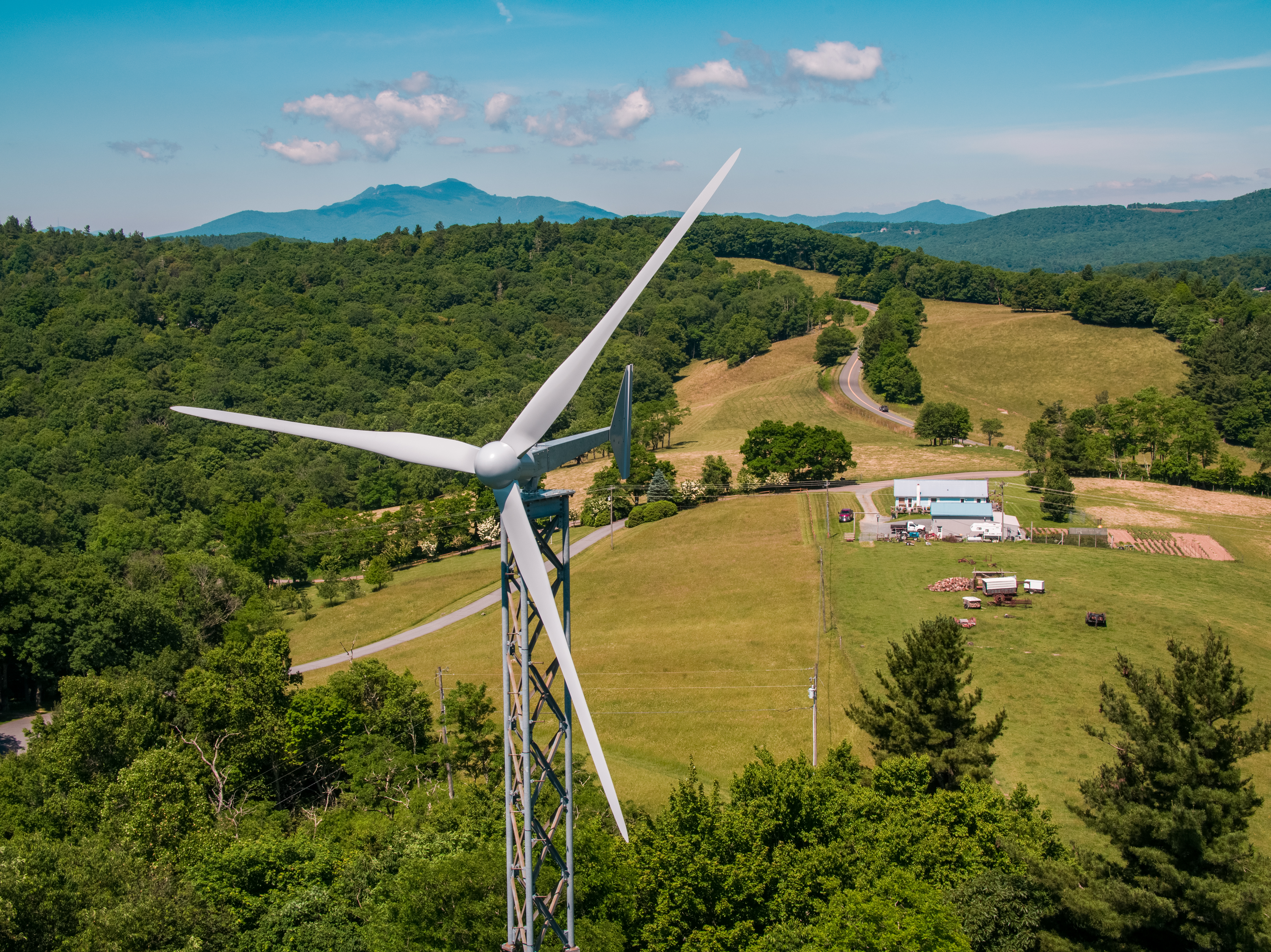 A small wind turbine in the foreground on a farm surrounded by forested mountains, with a country road winding away into the background