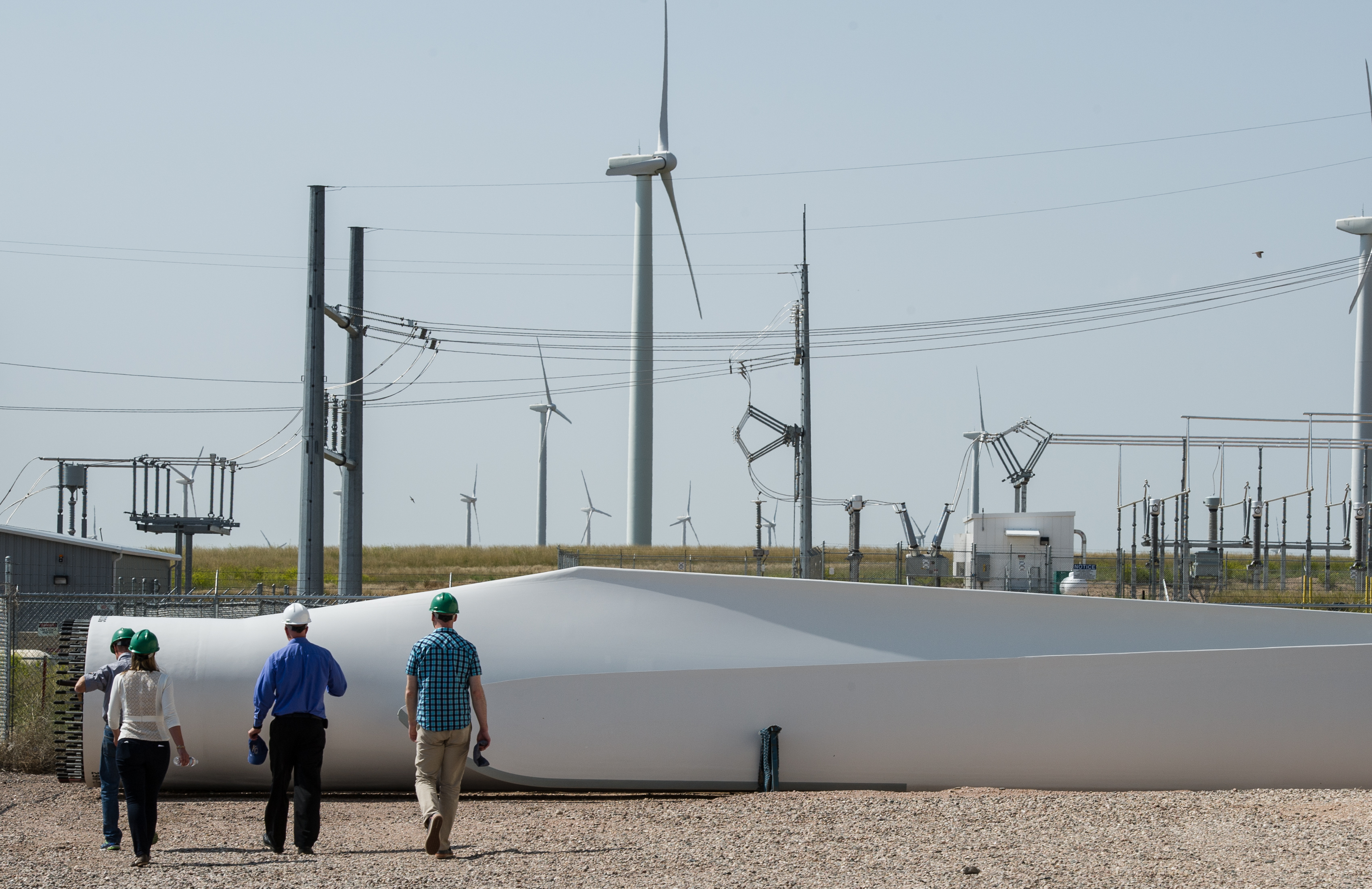 A group of people in hard hats walk toward a wind turbine blade lying on the ground. Power lines and more wind turbines are visible in the background