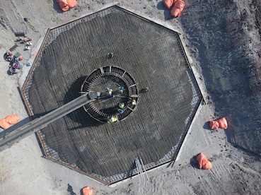 Overhead view of an octagonal base in a dug pit on which workers in protective attire walk.