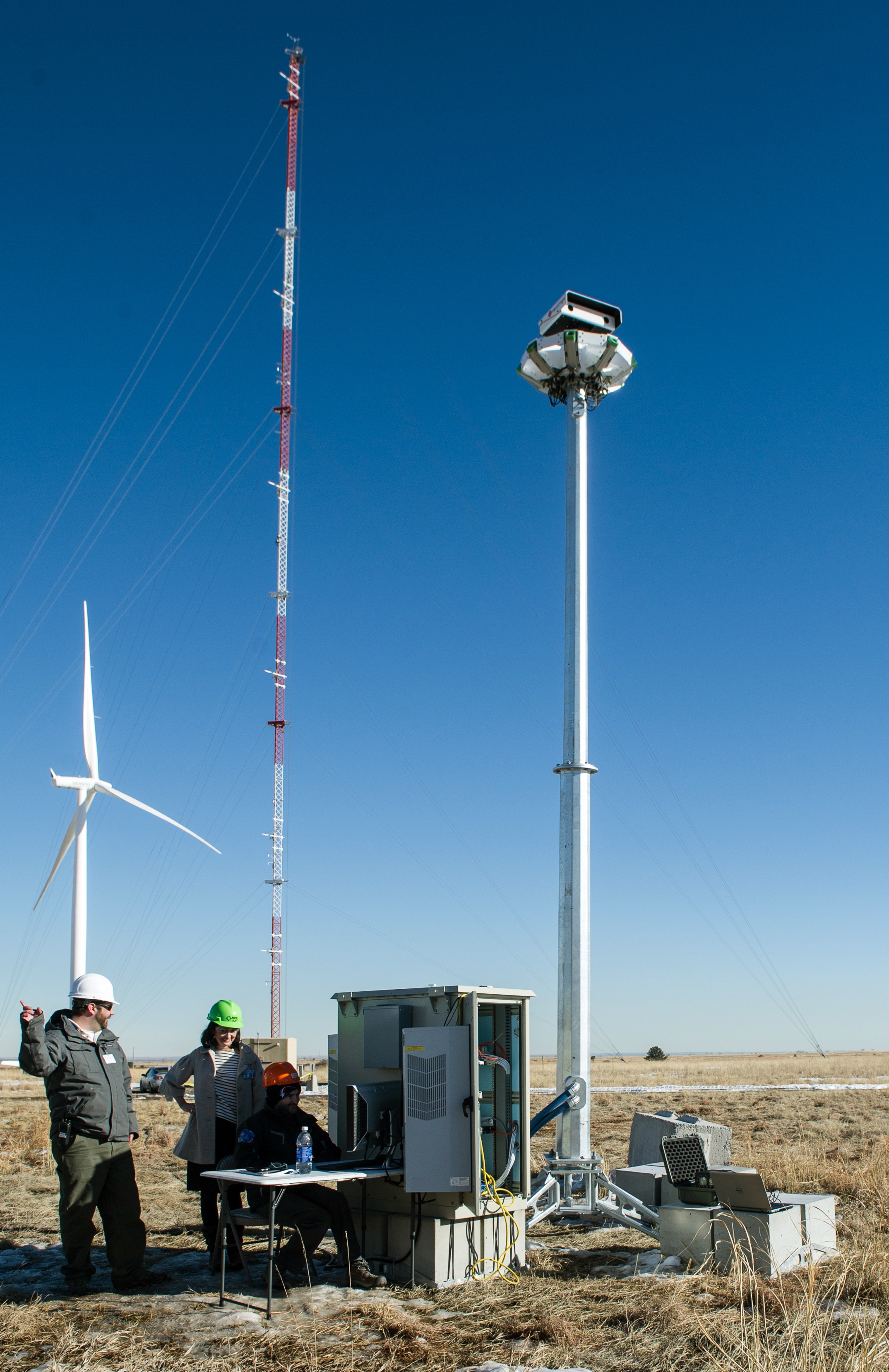 Two people in hard hats standing next to electrical devices on the ground and on a pole with a wind turbine in the background