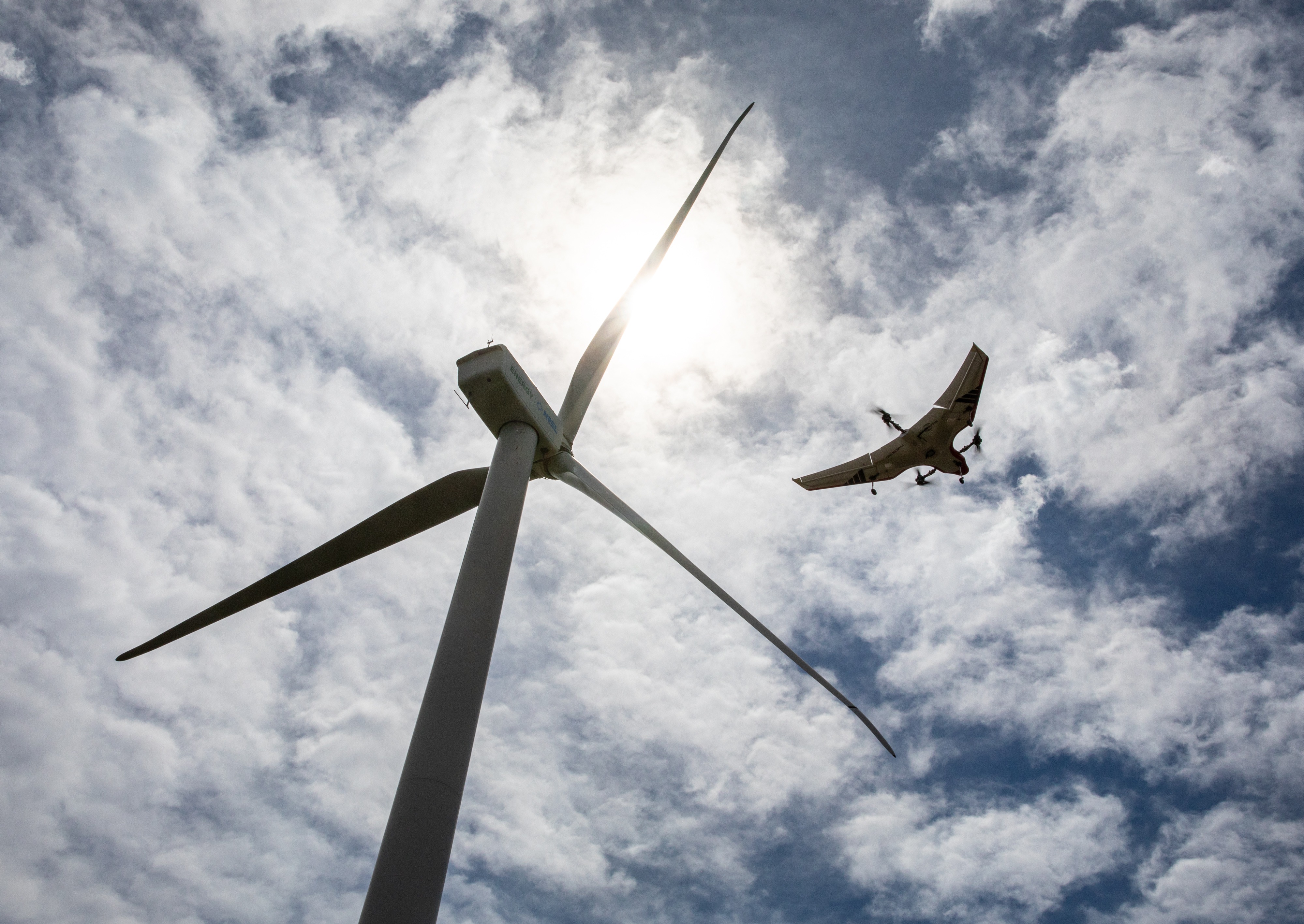 An unmanned aircraft flies over a wind turbine as viewed from below.