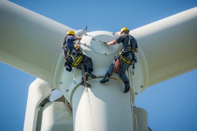 Two workers dangling from harnesses in front of a wind turbine rotor.