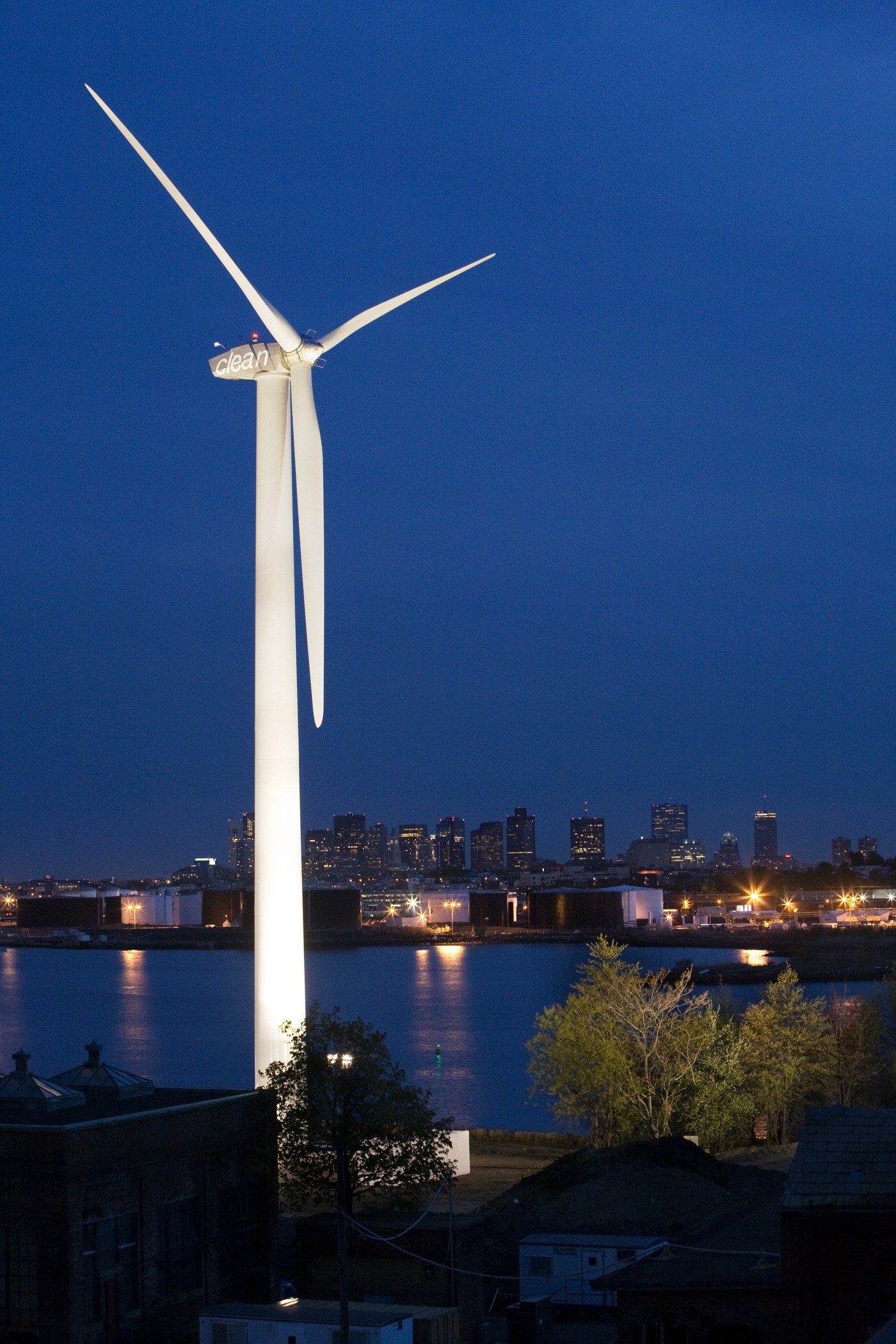 A wind turbine illuminated in front of a river across from a city at night